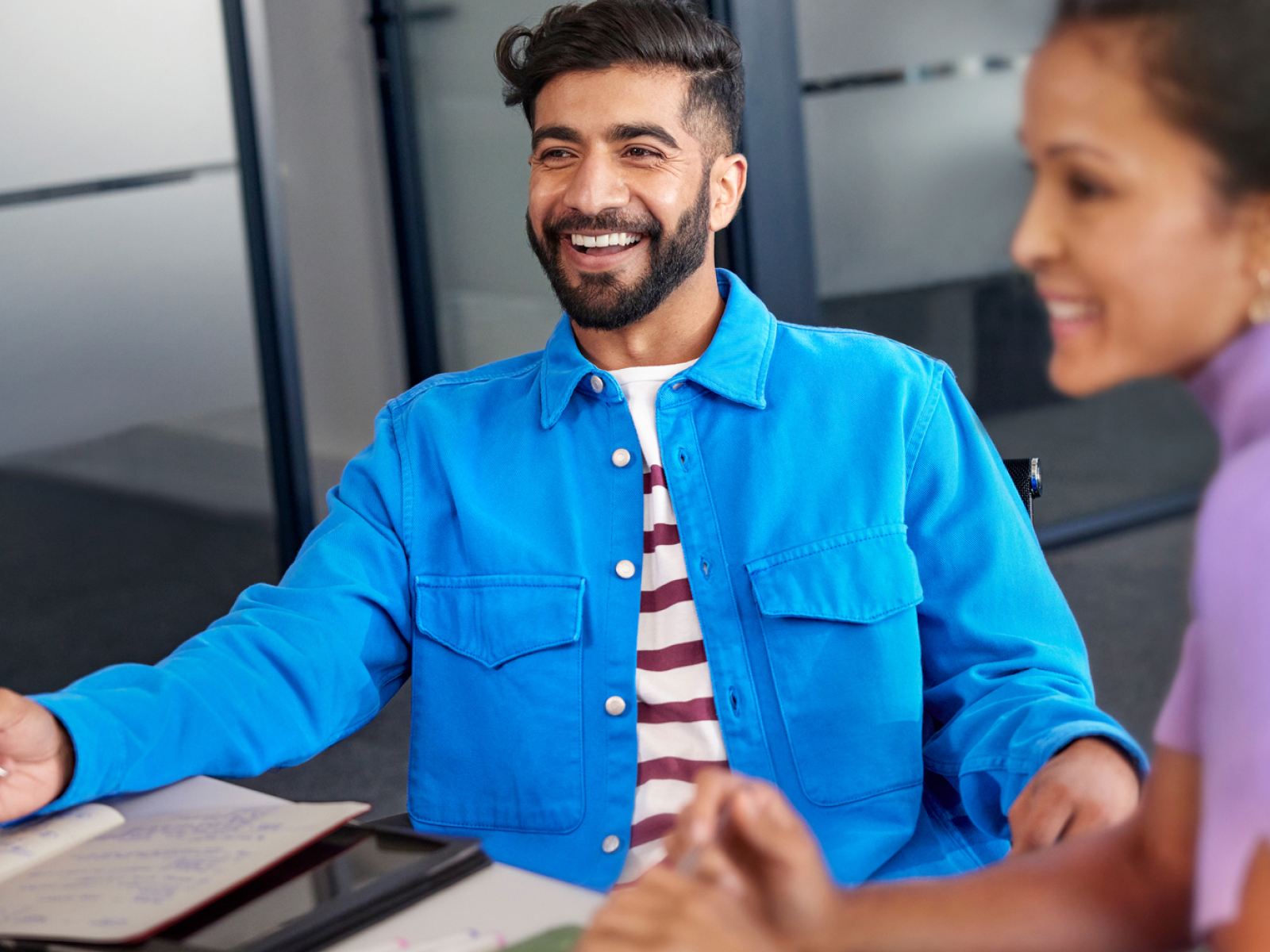 A man wearing a bright blue jacket and striped shirt smiles while sitting at a table with an open notebook, next to a woman who is also smiling. They appear to be in a modern office setting.
