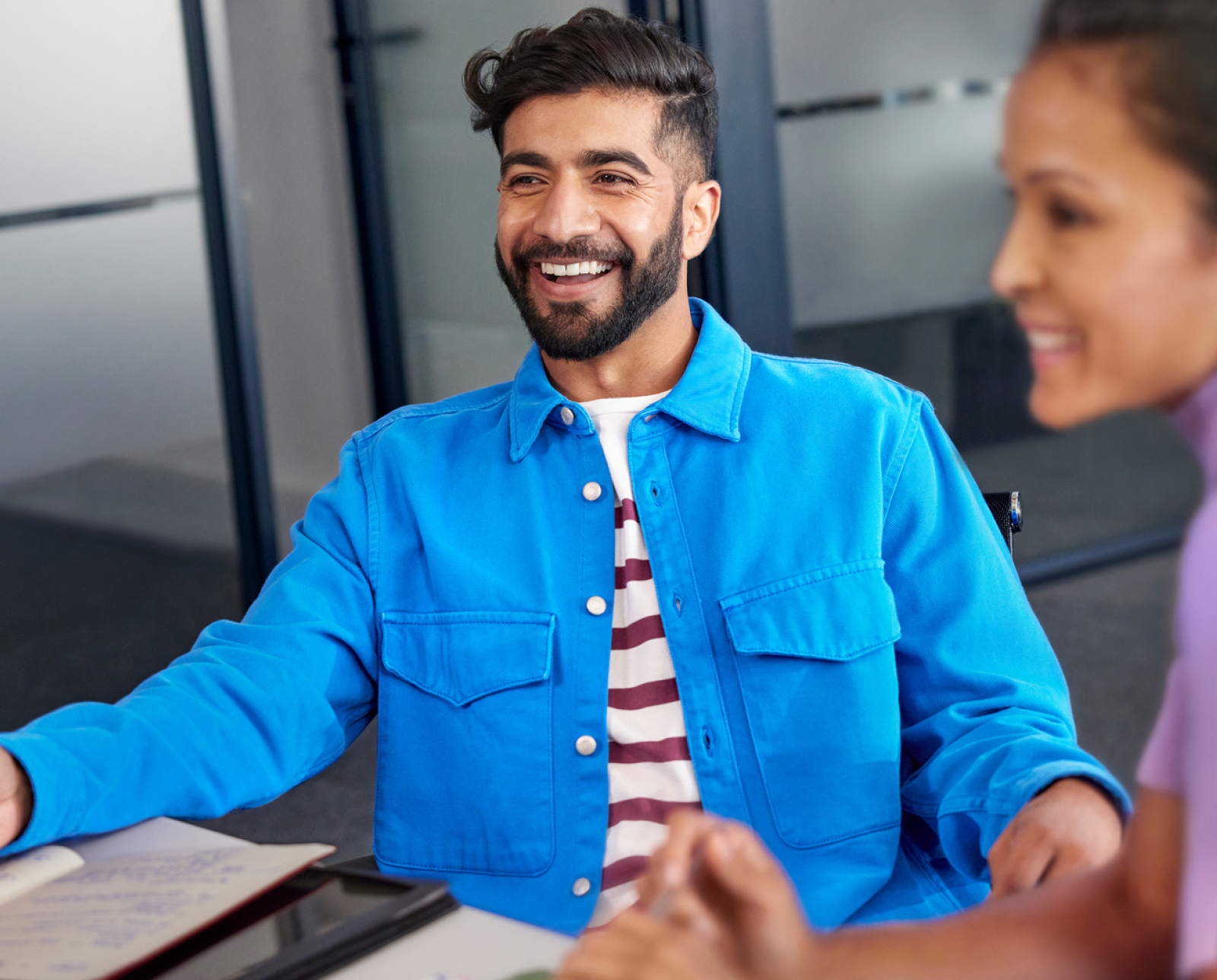 A man wearing a bright blue jacket and striped shirt smiles while sitting at a desk with a woman. They appear to be in a modern office, engaged in a meeting or discussion.