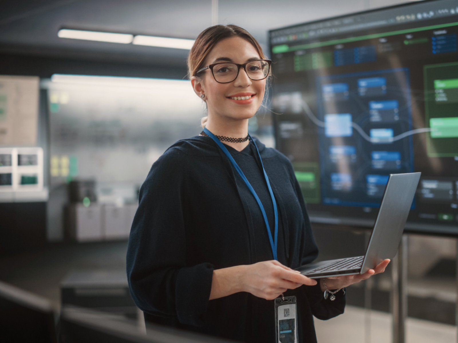A woman wearing glasses and a dark top holds a laptop and smiles in a modern office, standing in front of large screens displaying data and charts.