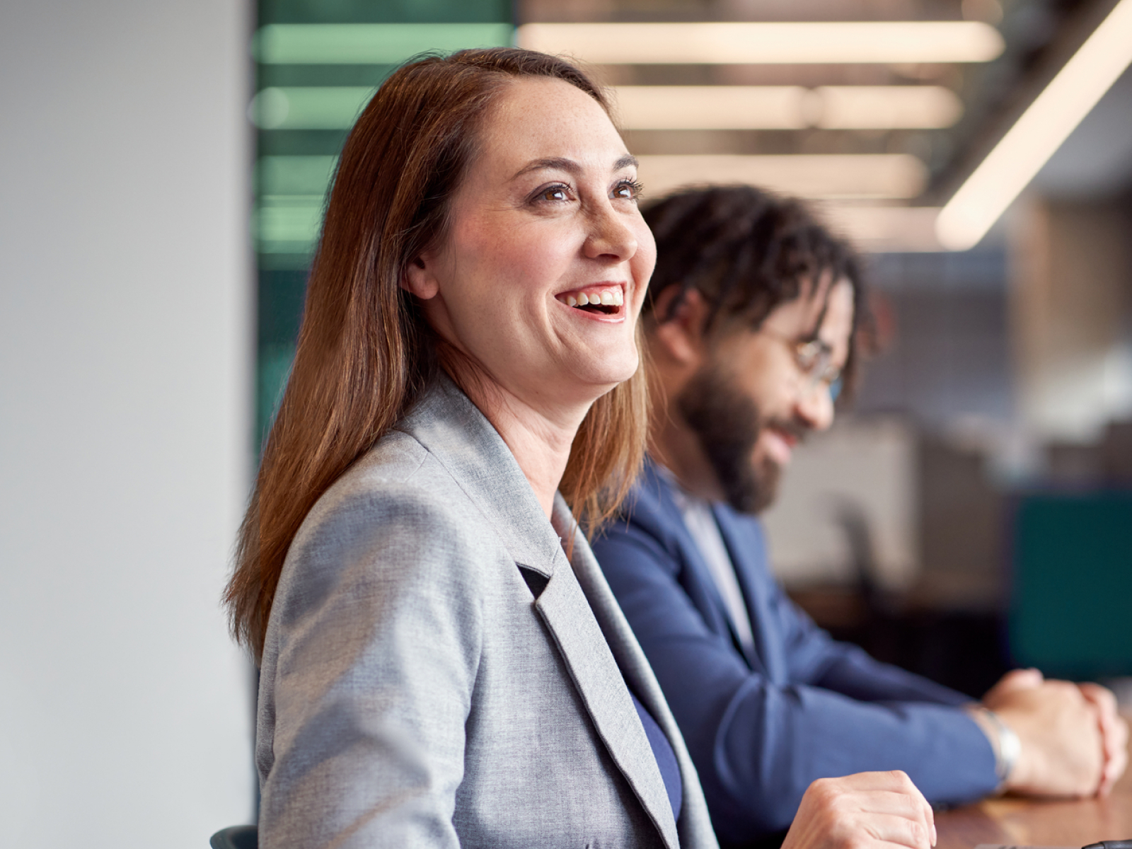 A woman in a gray blazer smiles and looks to the side while sitting at a desk next to a man in a blue suit, both in a modern office setting with blurred lights in the background.
