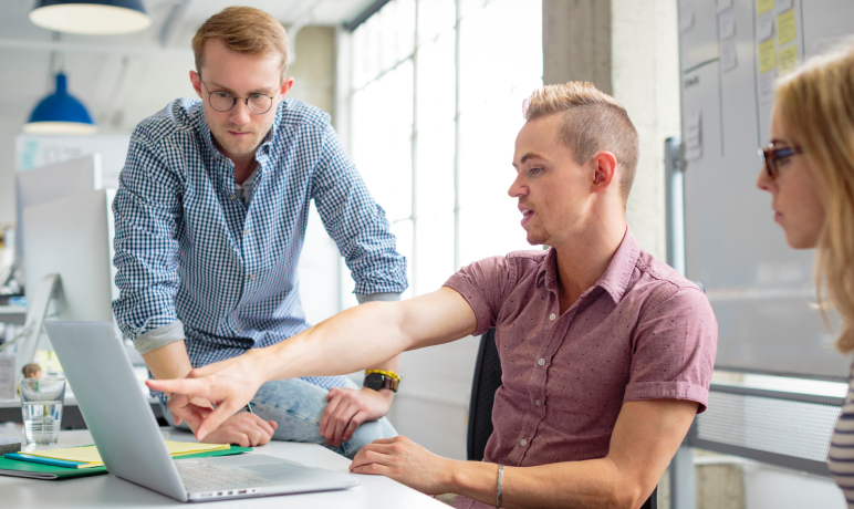 Three people in a modern office collaborate at a desk. One man sits and points at a laptop screen while another man and a woman, both standing or sitting nearby, listen and look at the screen attentively.