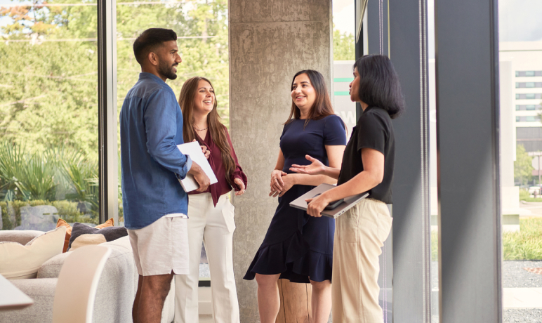 Four people stand in a modern office, smiling and talking together. Two hold laptops, and sunlight streams through large windows, highlighting the bright, open space and greenery outside.