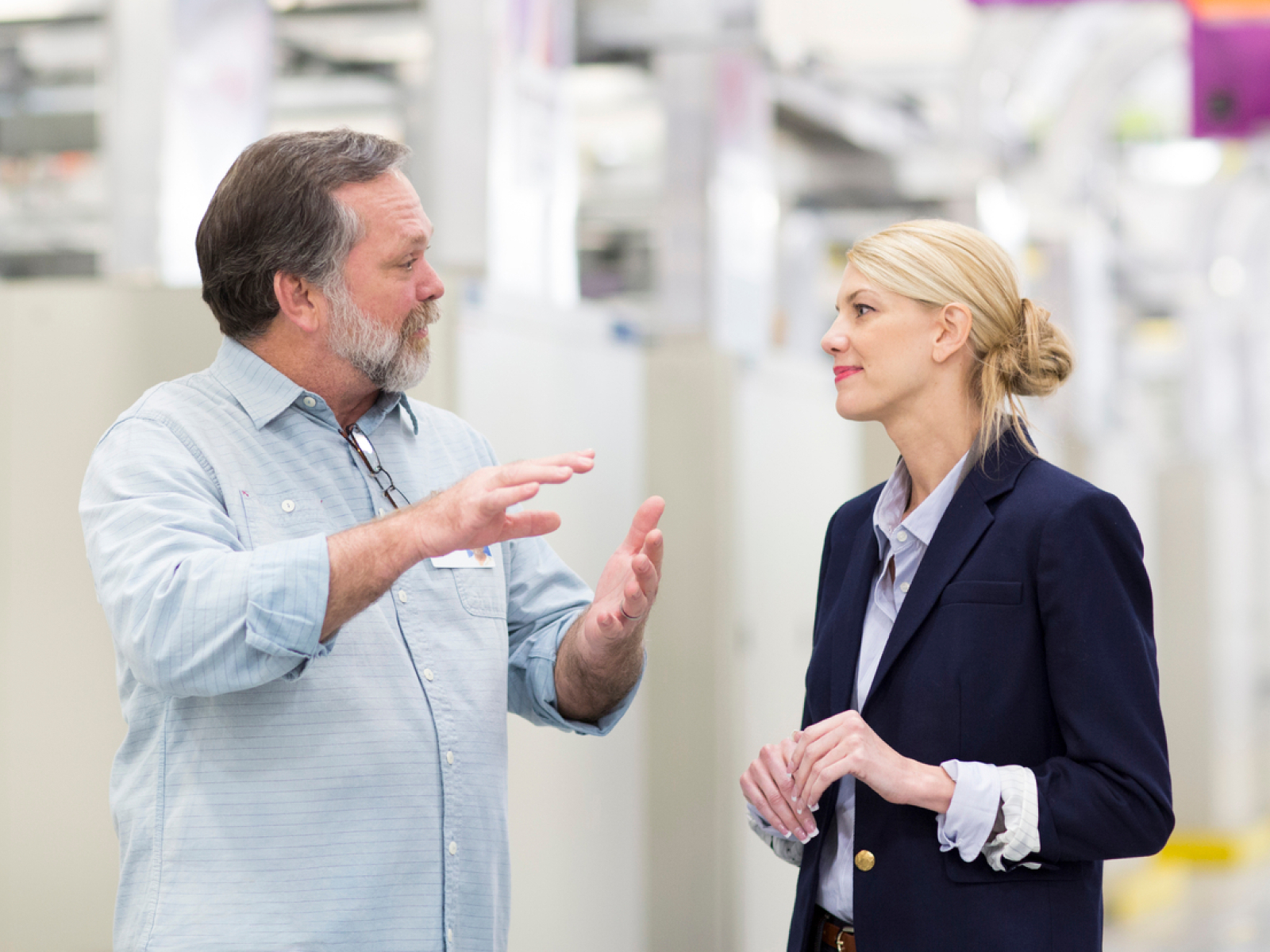 A man gesturing while speaking to a woman in a blazer. They are standing in a bright, modern industrial or office setting, appearing engaged in conversation.