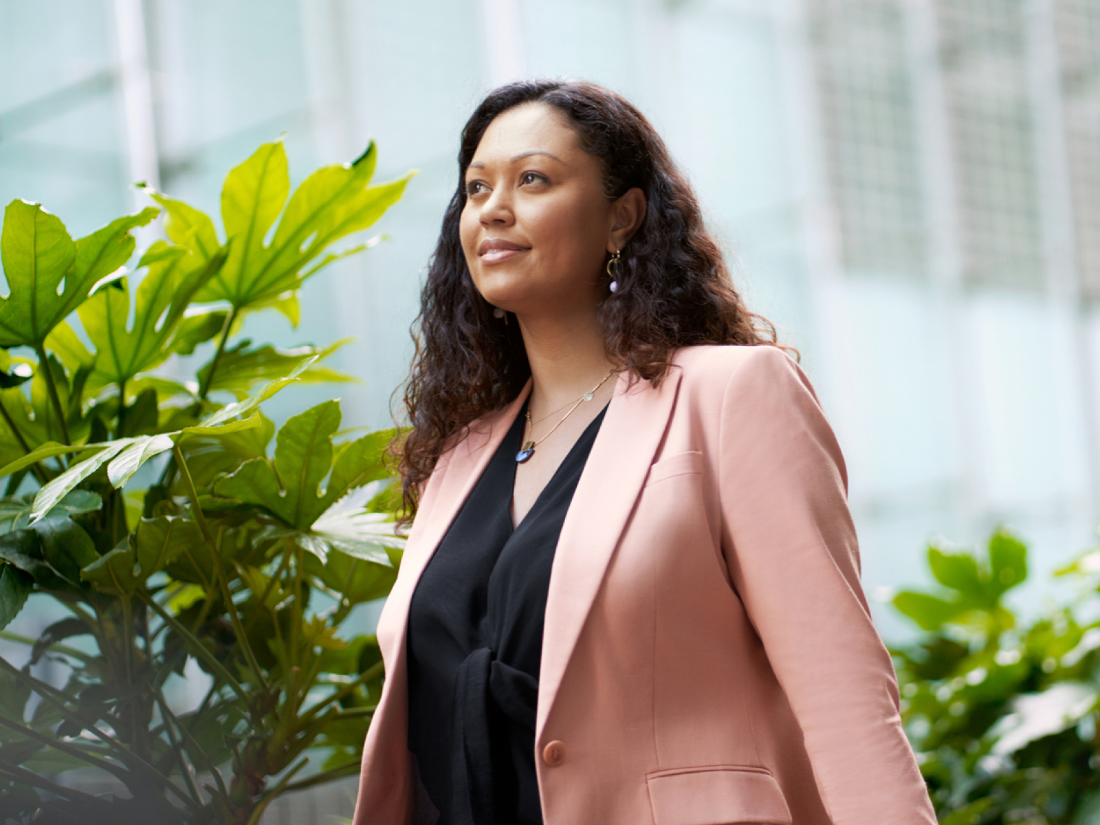 A woman with curly hair wearing a peach-colored blazer and black top stands outdoors near green plants, with a modern glass building in the background. She appears confident and thoughtful.