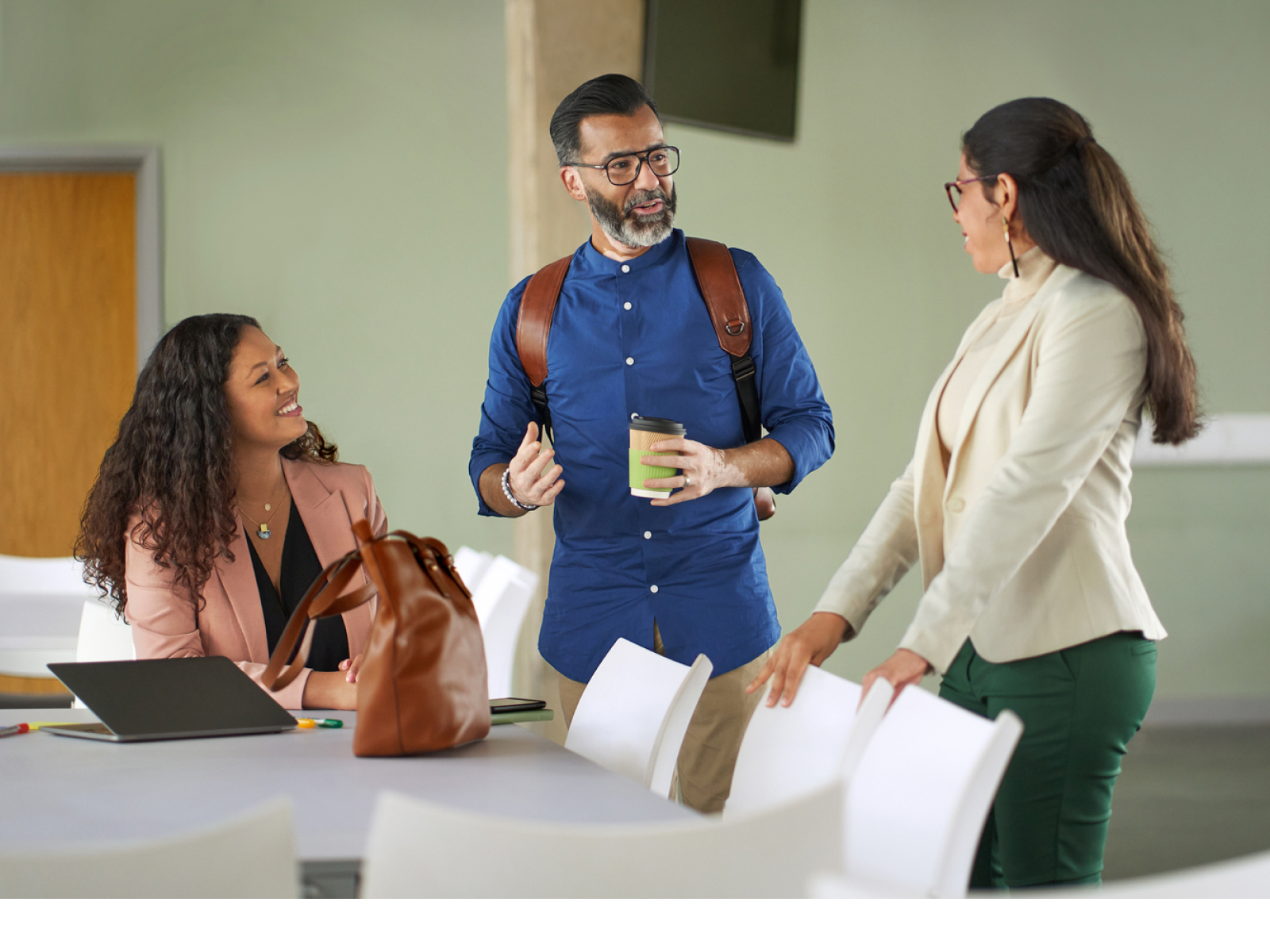 Three professionals have a discussion in an office setting. One woman sits at a table with a laptop, while a man with a backpack and a coffee cup talks to a standing woman in a white blazer.