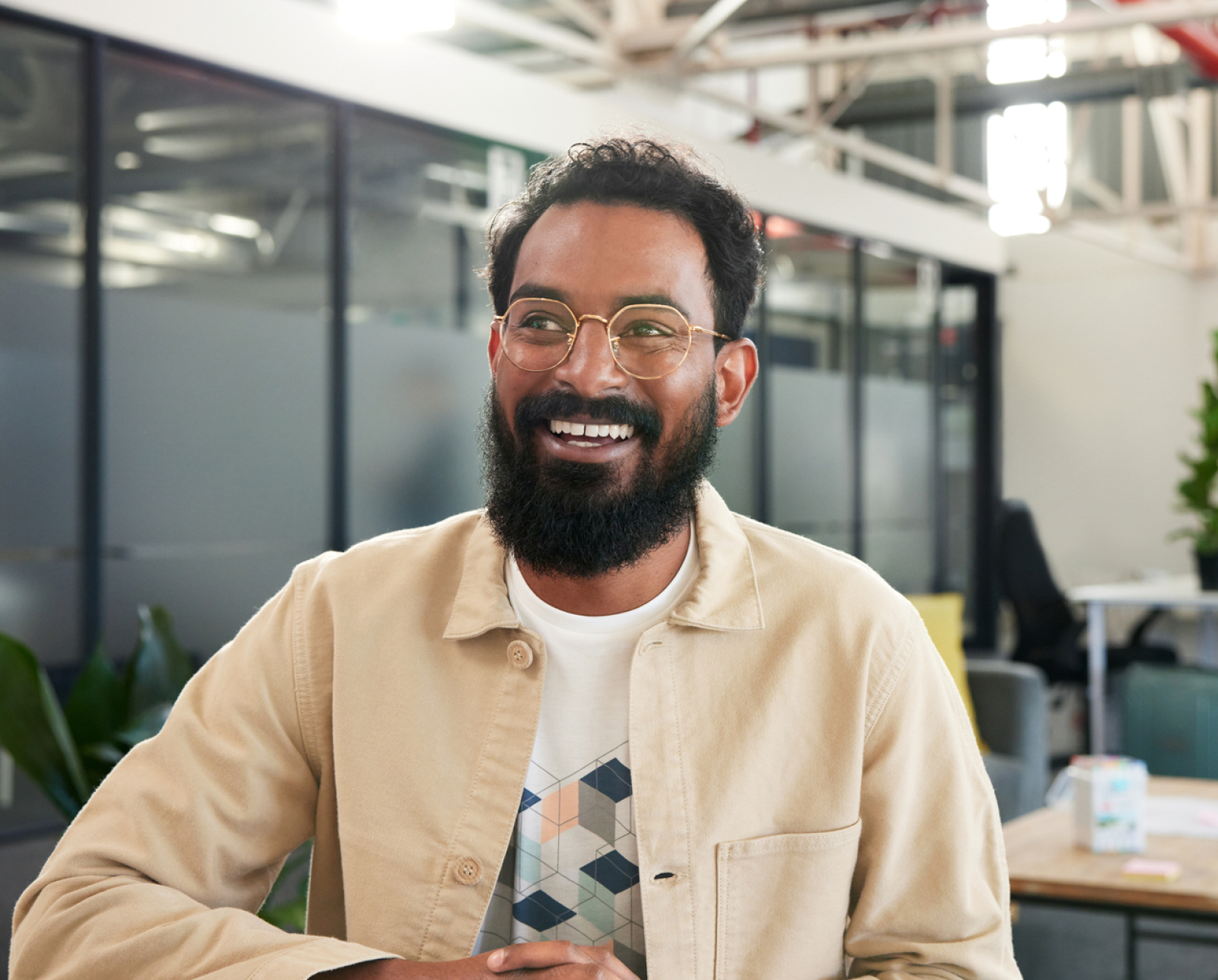A smiling man with glasses and a beard sits indoors in a modern office space, wearing a light-colored jacket over a patterned shirt. Glass walls and office furniture are visible in the background.