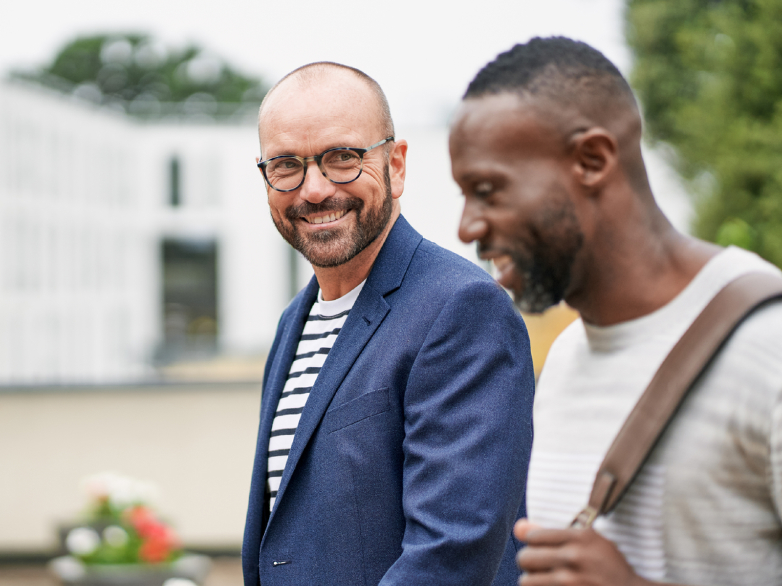 Two men are walking outdoors. One, wearing glasses and a blue blazer over a striped shirt, is smiling at the camera. The other, in a light sweater with a bag strap over his shoulder, is looking down and smiling.