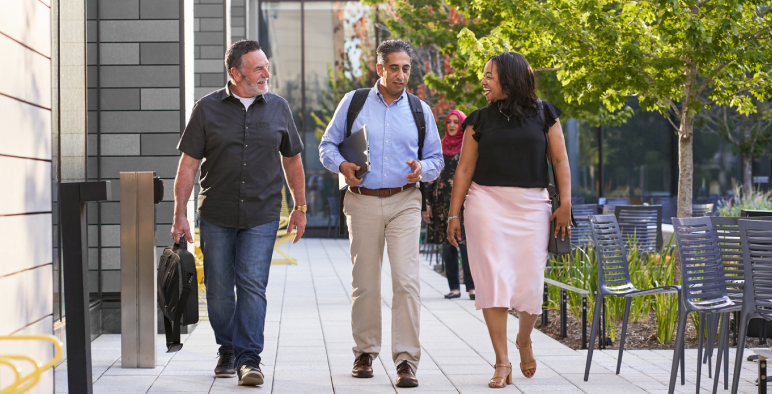Three people walk together outside on a sunny day, smiling and talking. They are dressed in business casual attire and passing by outdoor seating and trees on a paved path.