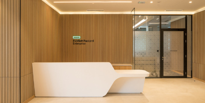 A modern office reception area with a sleek white desk, light wood-paneled walls, and glass doors. The Hewlett Packard Enterprise logo is on the wall behind the desk. The space is bright and minimalist.