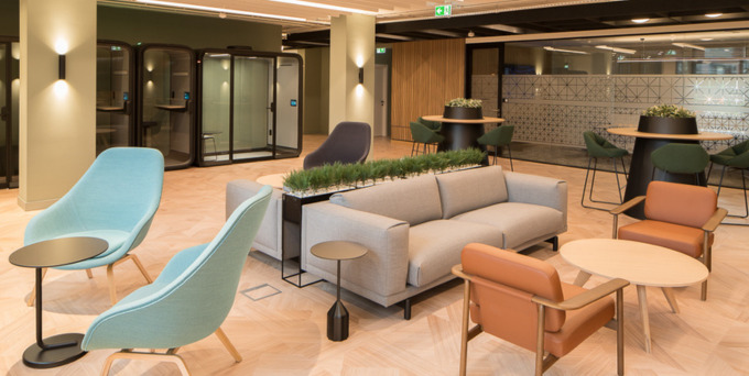 A modern lounge area with pastel blue and tan armchairs, a light gray sofa, small round tables, a planter divider, and booths in the background. The room has wood flooring and soft, ambient lighting.