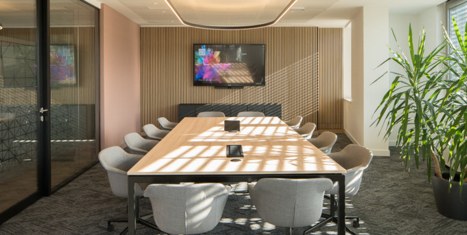 A modern conference room with a long wooden table surrounded by grey chairs, a flat-screen TV mounted on a wood-paneled wall, large windows letting in sunlight, and a tall green plant in the corner.