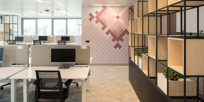 Modern open-plan office with rows of desks and computers, ergonomic chairs, a geometric patterned accent wall, and shelves with plants and storage boxes. Large windows let in natural light.