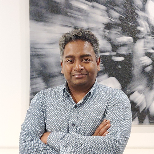 A man with tan skin standing with arms crossed in front of a blurred black-and-white background. He has a soft smile not showing teeth, and the monochromatic backdrop adds a dramatic, high-contrast visual effect.