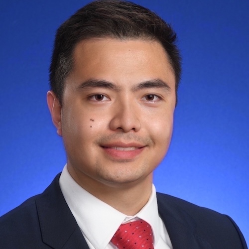 Professional headshot of a male Lilly employee against a blue gradient background. The individual has light tan skin, dark hair and is wearing a dark suit jacket, white dress shirt, and patterned red tie, facing the camera in a studio-style portrait with shoulders visible.
