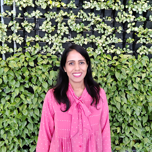 A woman with tan skin and black hair wearing a pink top, standing in front of a lush green wall covered with small white flowers. She is smiling with her teeth, and the vibrant foliage creates a natural, serene atmosphere.