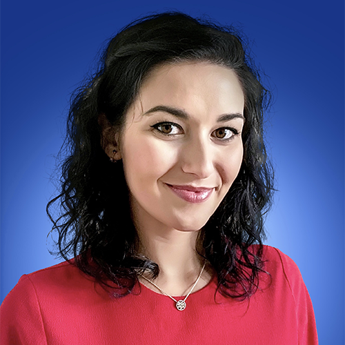 A woman wearing a red dress and dark wavy hair sits against a solid blue background. She is similing without teeth looking directly at the camera.