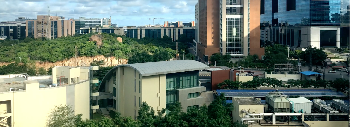 Panoramic view of an urban landscape featuring a mix of modern architecture and greenery. In the foreground, a building with a curved roof and large glass windows is surrounded by trees and adjacent structures, including industrial equipment. The background showcases several tall office buildings with reflective glass facades, and construction cranes are visible in the distance under a partly cloudy sky.