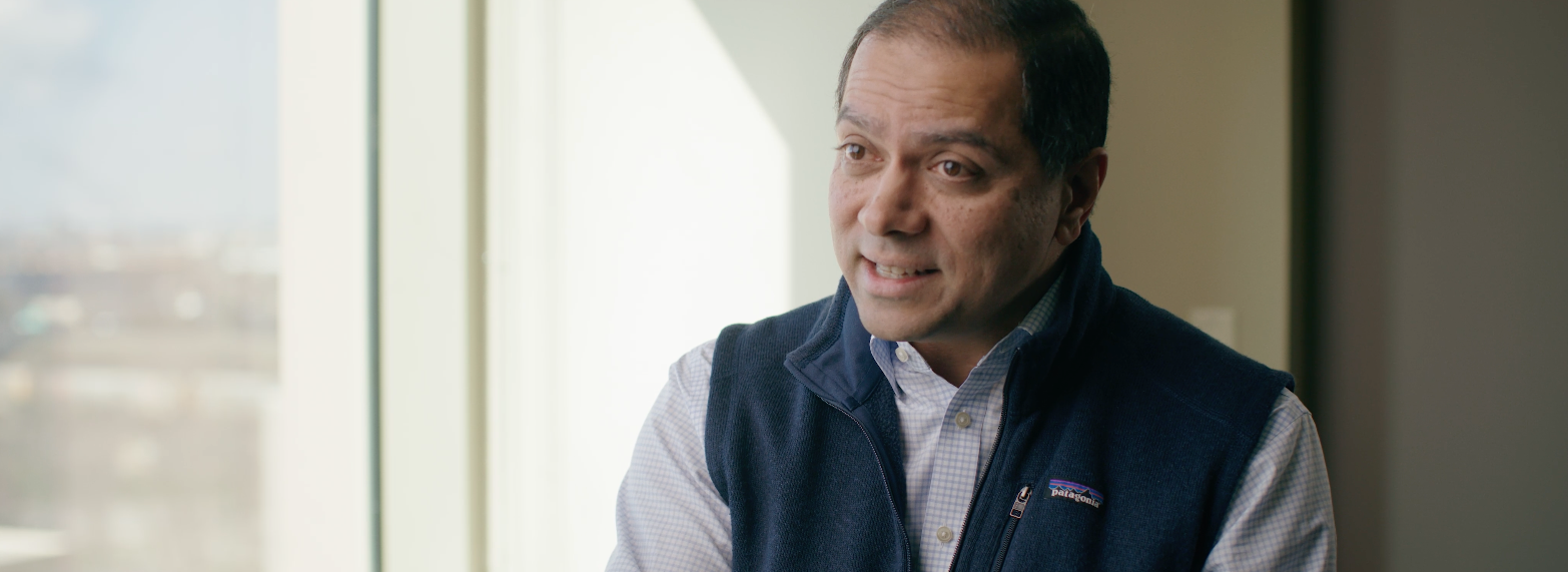 A man with tan skin wearing a dark blue quilted vest over a light-colored, checkered button-down shirt stands indoors near a large window. Natural daylight streams through the window, softly illuminating the scene. The background reveals an out-of-focus urban environment with buildings and greenery visible through the glass.