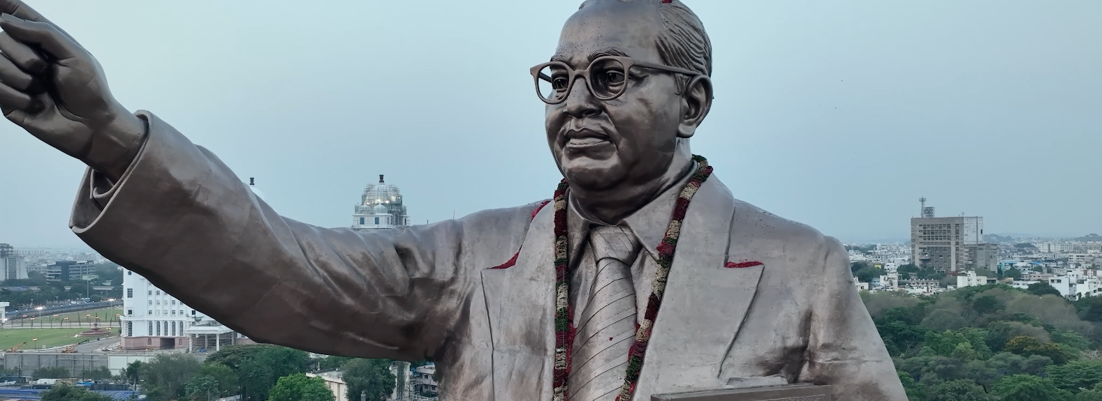Statue of a person wearing glasses, a suit, and a tie, with one arm raised and pointing forward. The statue’s face is intentionally blurred for privacy. Set in an urban environment, the background includes buildings, trees, and an open plaza area, suggesting a public or civic space.