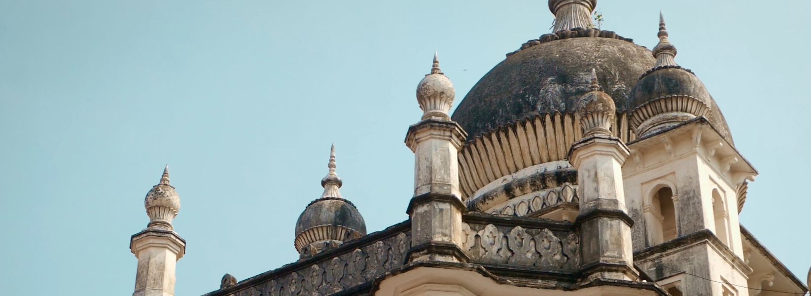 Architectural detail of a historic stone structure featuring a large central dome flanked by smaller domes and ornate spires. The building showcases intricate carvings and weathered textures, suggesting age and craftsmanship. Set against a clear blue sky, the image highlights the grandeur and symmetry of the design.