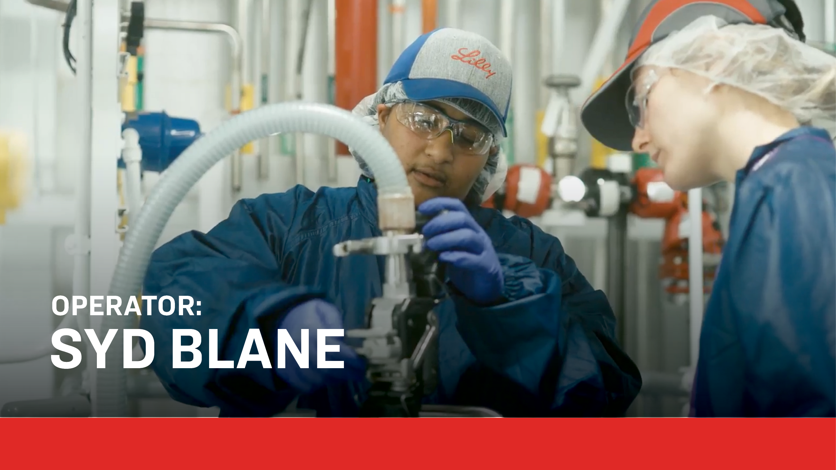 Two operators wearing gloves, goggles, and hair coverings collaborate while adjusting machinery in a pharmaceutical production environment. Text overlay reads: “Operator: Syd Blane,” highlighting hands-on roles supporting medicine manufacturing.