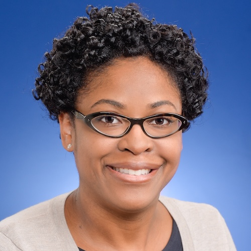 Professional headshot of a female Lilly employee against a blue gradient background. The individual has brown skin, dark hair, and is wearing dark-framed glasses, a dark top, and a light cardigan, facing the camera in a studio-style portrait with shoulders visible.