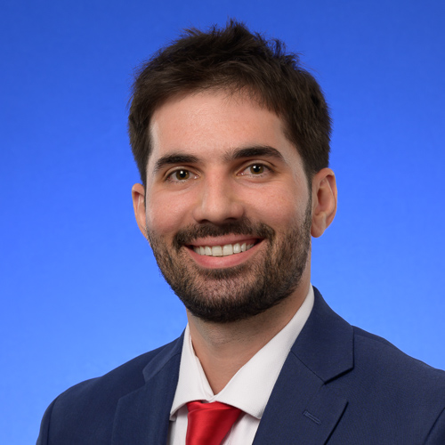 A man in a suit and tie stands against a solid blue background. He is similing with teeth and has dark facial hair looking directly at the camera.