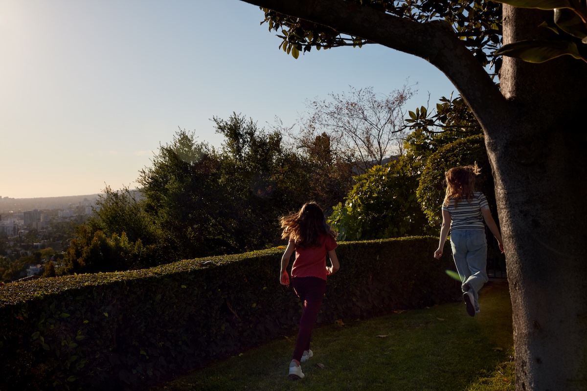 Two children with long brown hair run along a grassy path beside a hedge and large tree overlooking a city skyline. Warm sunlight shines across the outdoor scene, representing family wellbeing, flexibility, and support for employees' personal lives.
