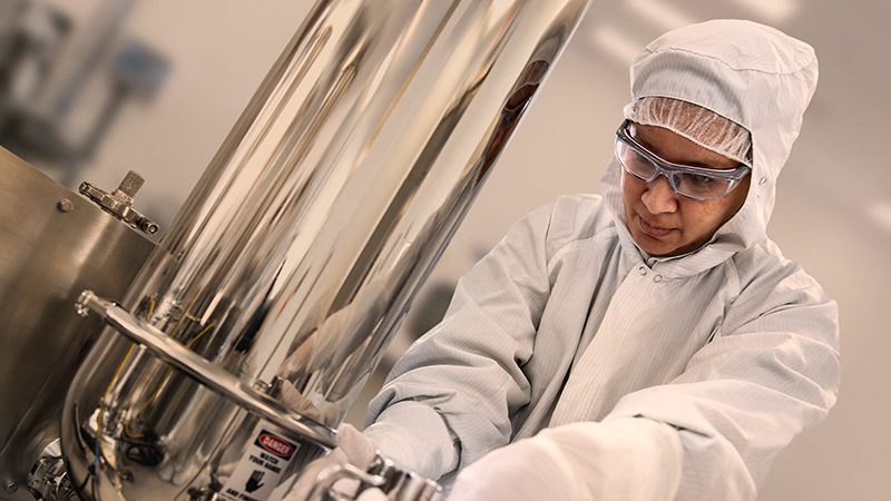 Individual wearing cleanroom protective clothing and safety glasses adjusting stainless steel manufacturing equipment in a pharmaceutical production environment.