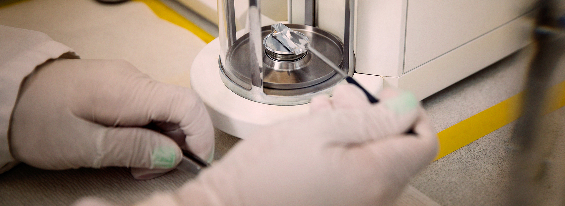 Close-up of gloved hands using a precision balance scale in a laboratory, with a small container or vial visible in the background