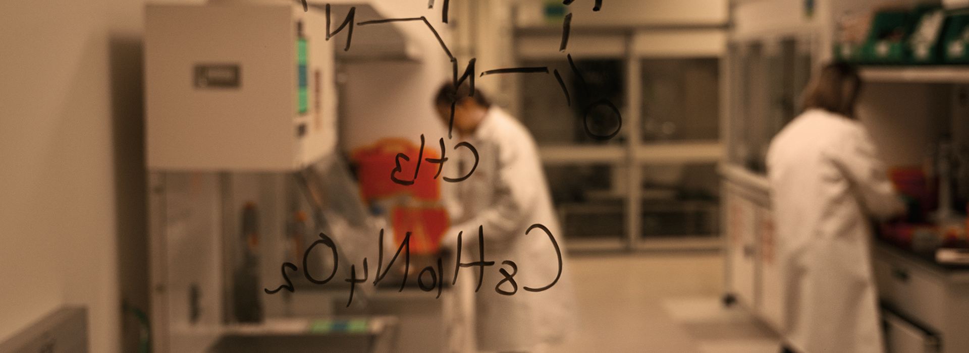Two individuals in lab coats working in a laboratory. A transparent surface in the foreground displays a chemical structure with the text: CH₃, CH₃, C₆H₄N₂O₂.