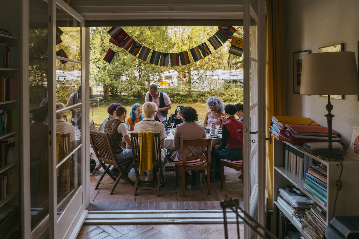 A diverse group of people gather around an outdoor table in a garden setting, viewed through open doors from inside a home. Colorful flags hang above the group as they share a meal and conversation, highlighting inclusion, community, belonging, and employee wellbeing.