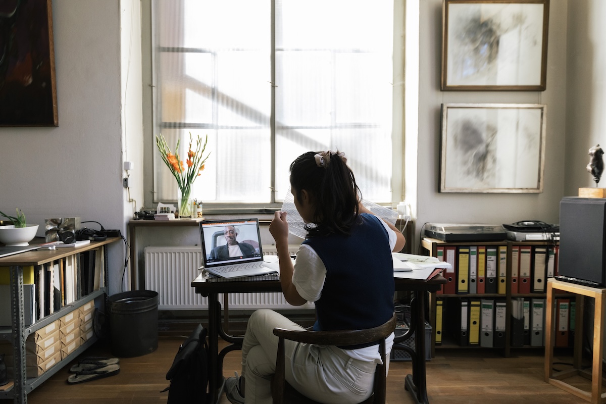 Person with dark hair tied back sits at a desk in a home office during a video call on a laptop. Documents, binders, and work materials surround the workspace, illustrating workplace flexibility, remote work, and support for balancing professional and personal responsibilities.