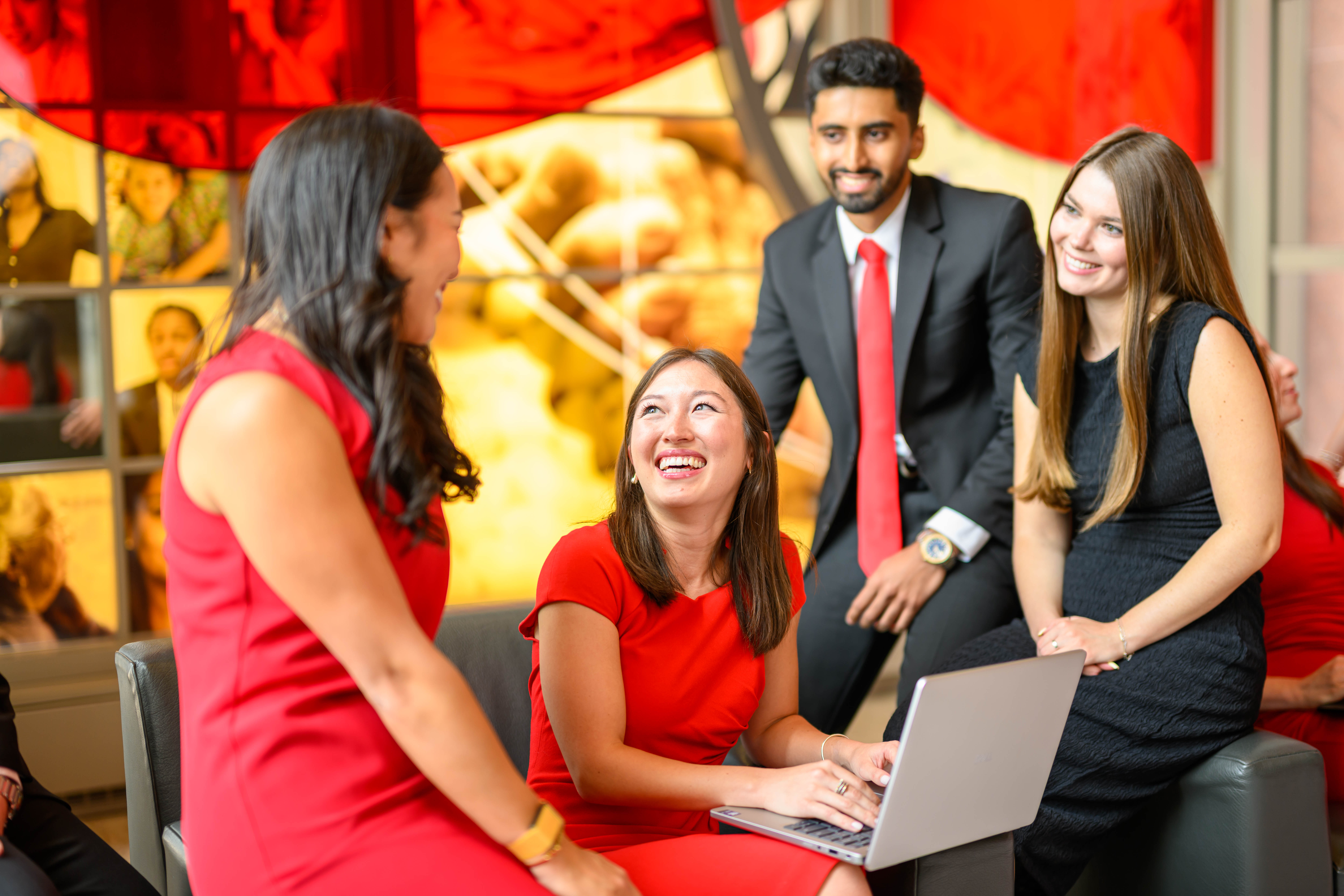 Four people—three women and one man—gathered around a laptop, with a red and yellow abstract design in the background. Faces are blurred for privacy.