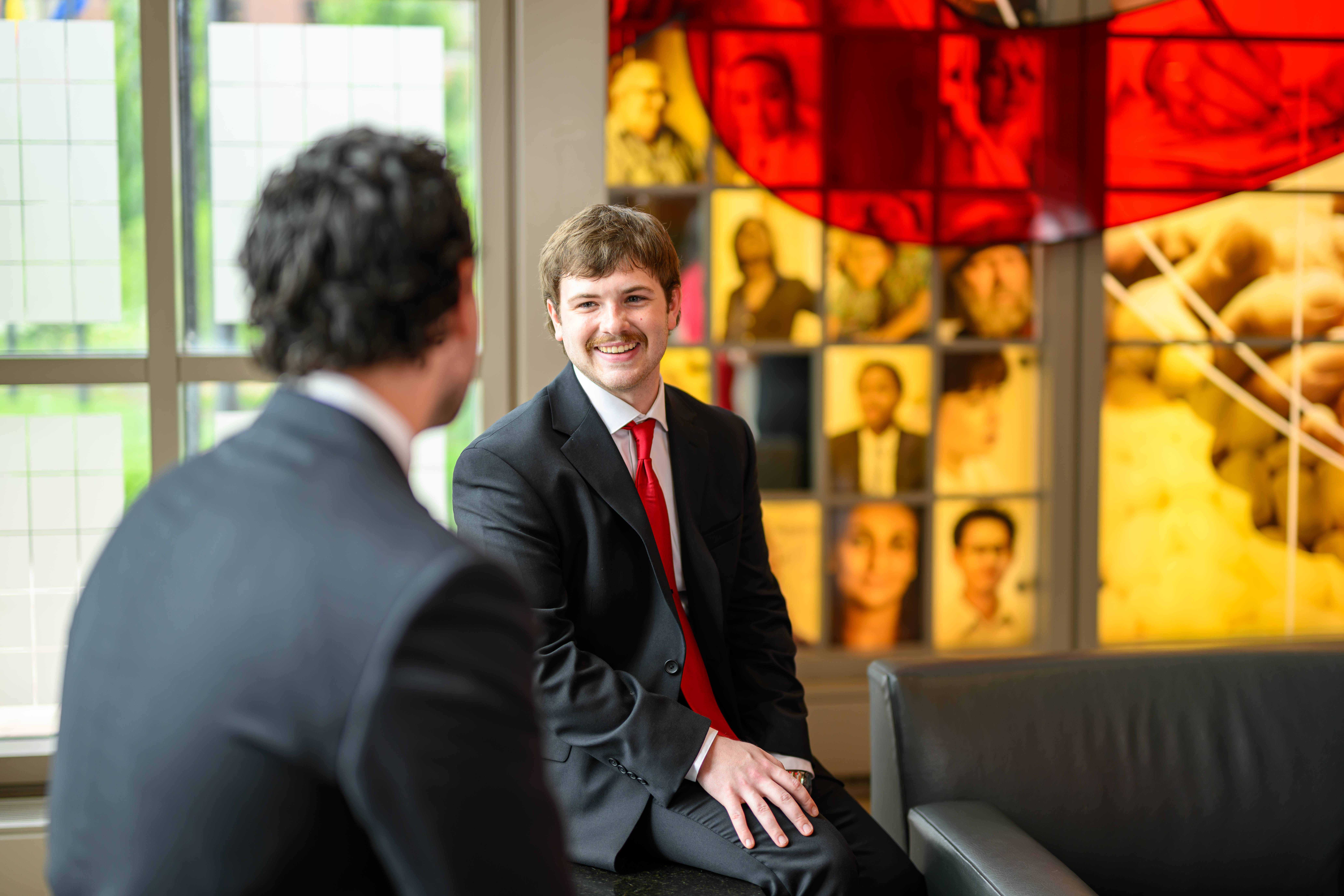 Two individuals in business attire seated and conversing in a professional setting, with a large, colorful stained glass window depicting various people in the background.