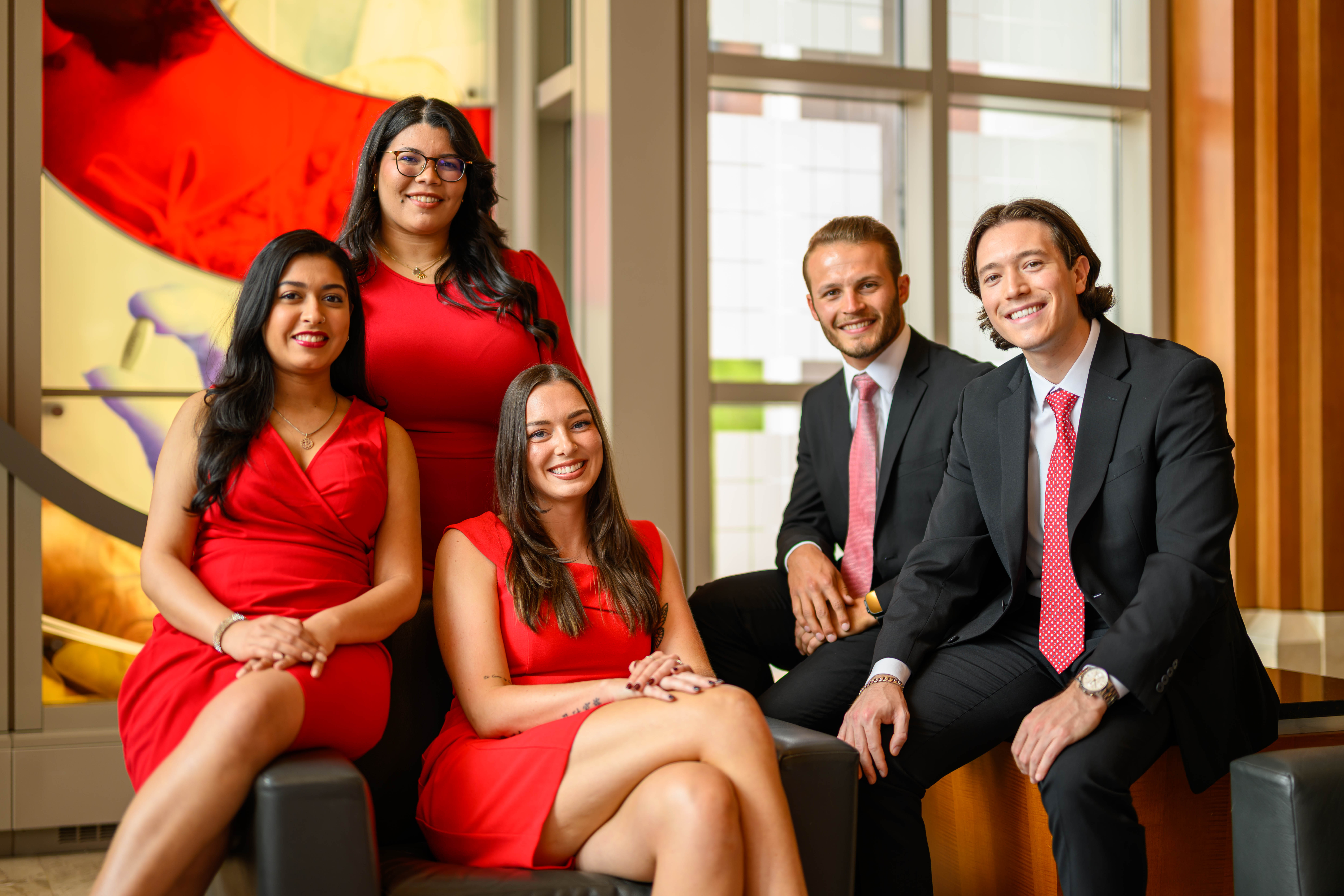 Five individuals—three women in red dresses and two men in black suits with white shirts and red ties—posed in a formal setting with a colorful abstract design on the left and large windows on the right.