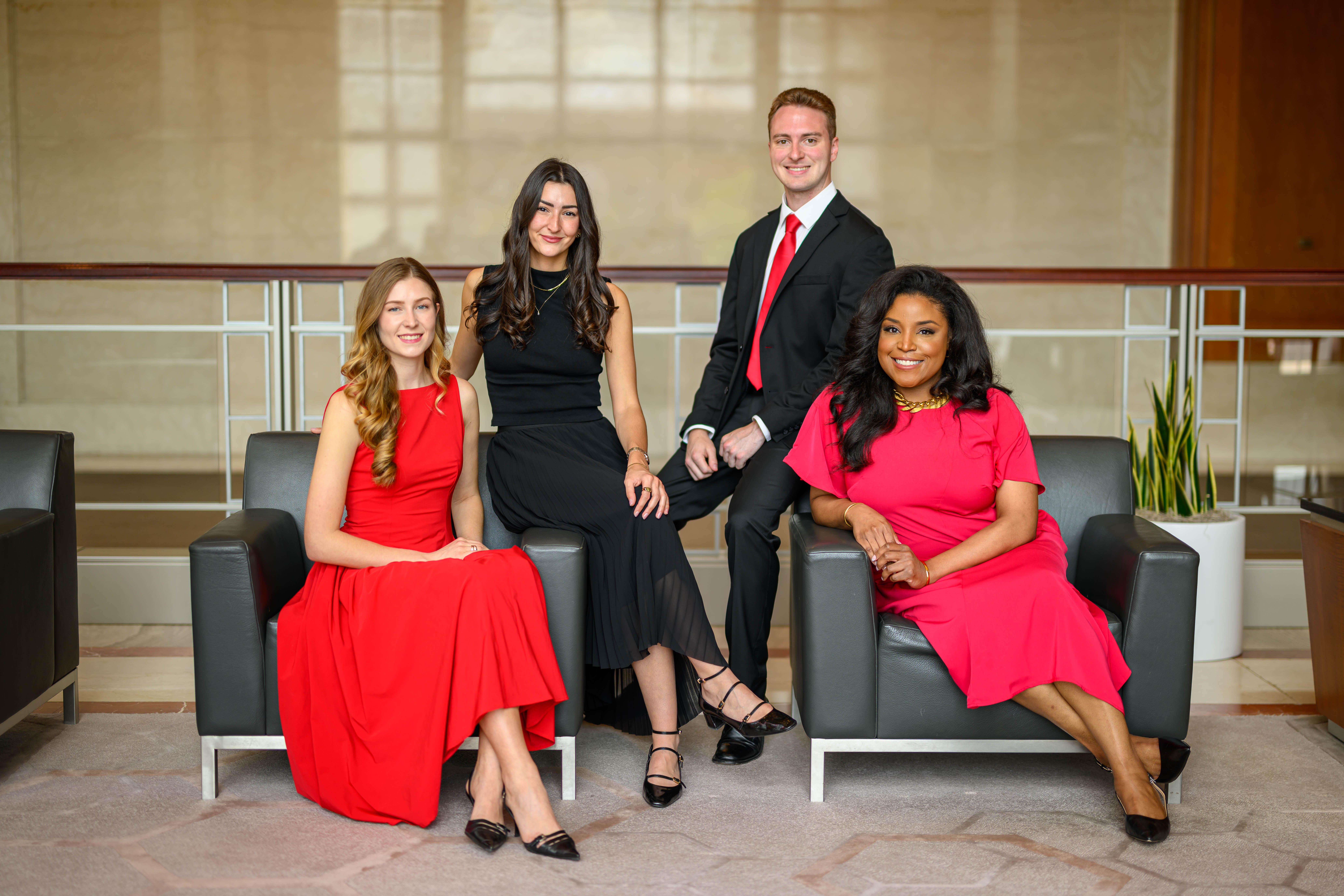 Four individuals seated on two black chairs in a formal setting. Three women are dressed in red and black dresses, and one man is wearing a black suit with a red tie.