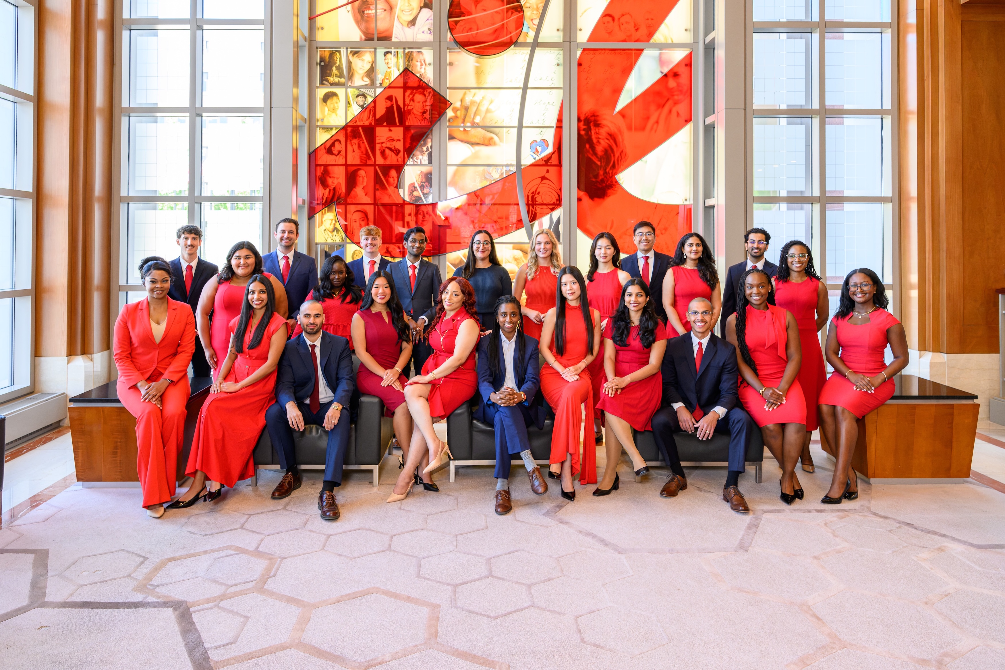 Large group of professionals gathered for a team photo in a bright corporate atrium. Employees wearing coordinated red and navy business attire are positioned in front of a large decorative wall display showcasing images and graphics, highlighting a collaborative workplace culture.