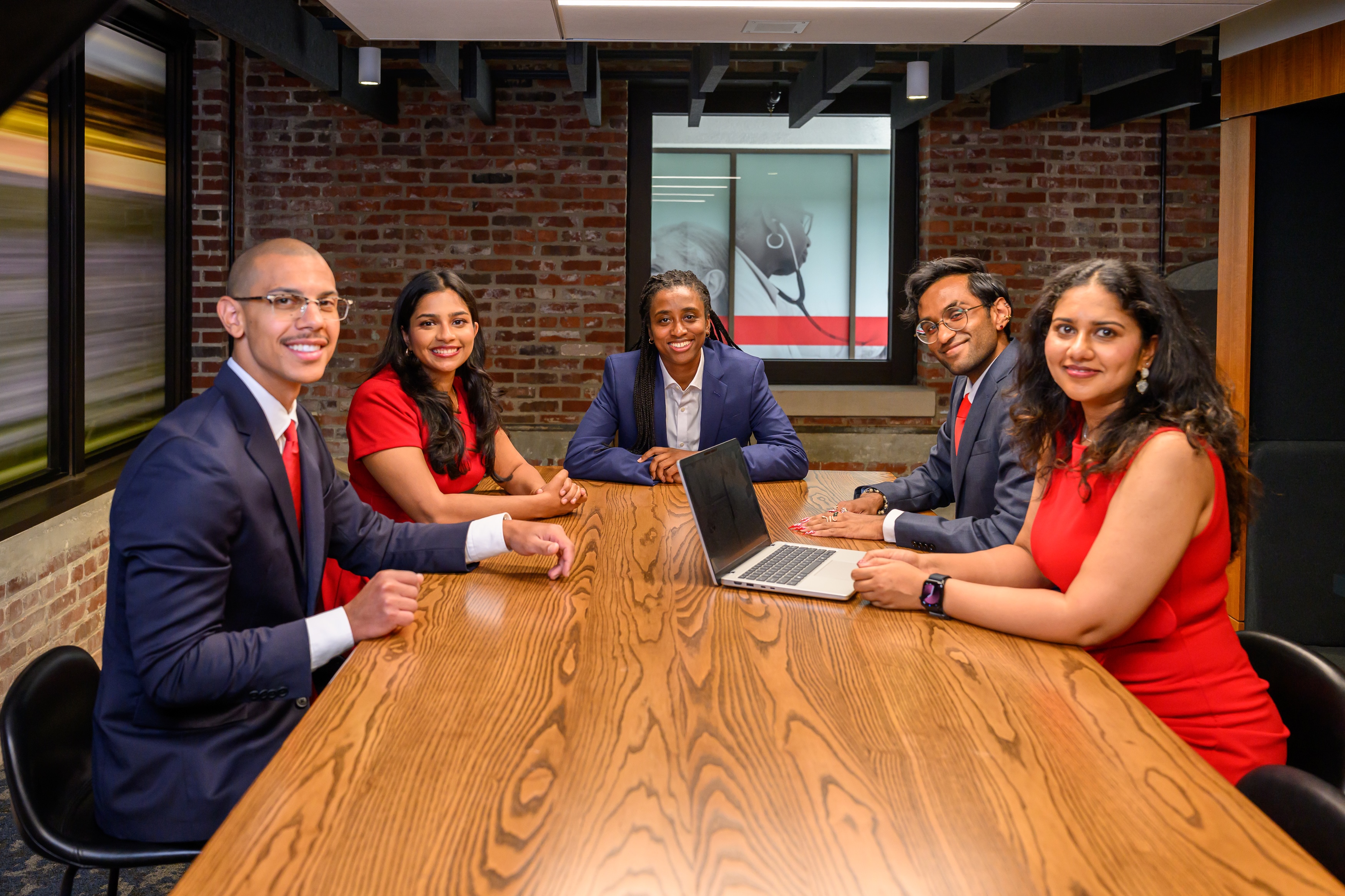 Five professionals seated around a conference table in a modern office meeting room, collaborating during a discussion with an open laptop. Exposed brick walls, large windows, and healthcare-focused artwork create a professional workplace setting.