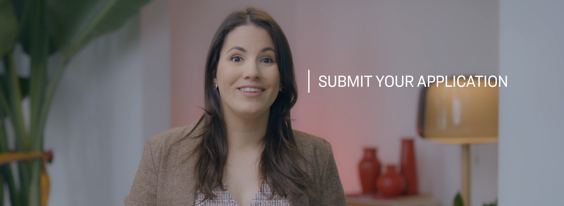 Woman with long dark brown hair wearing a brown blazer and patterned blouse, seated indoors facing the camera. A large green plant is visible on the left, with red decorative vases and a table lamp in the background. White text on the right side of the image reads, "Submit Your Application."