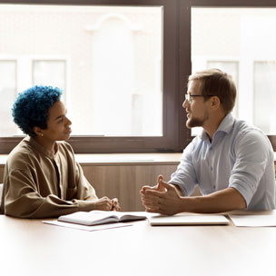 Two people sit at a table having a serious conversation, with open notebooks in front of them and large windows in the background.