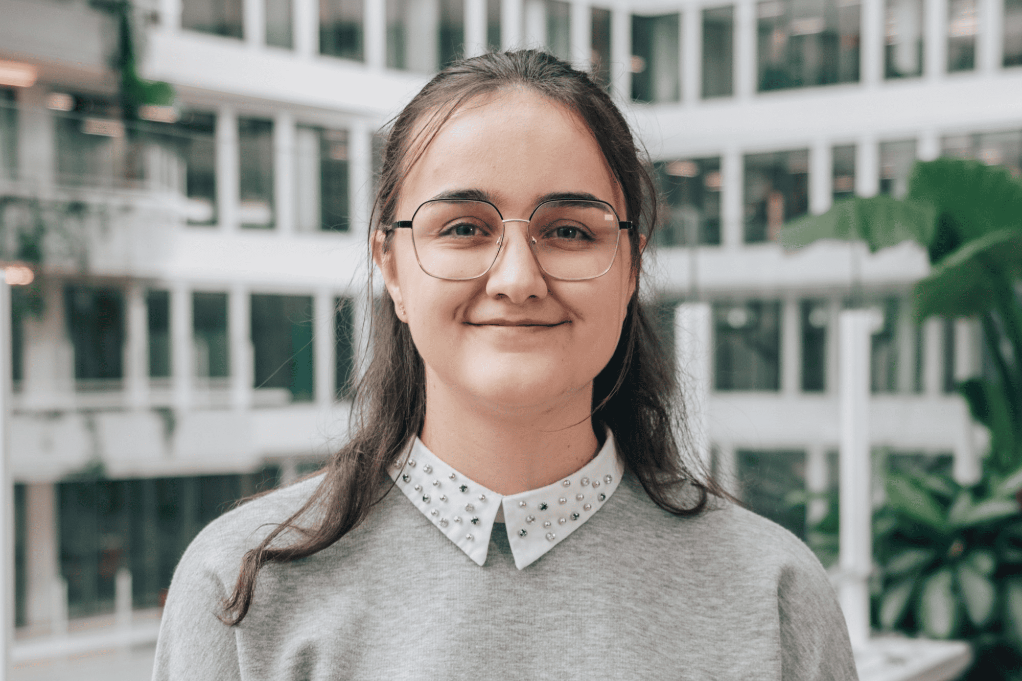A bright, smiling portrait of Claire, a student intern, wearing a grey shirt in a modern glass office atrium.