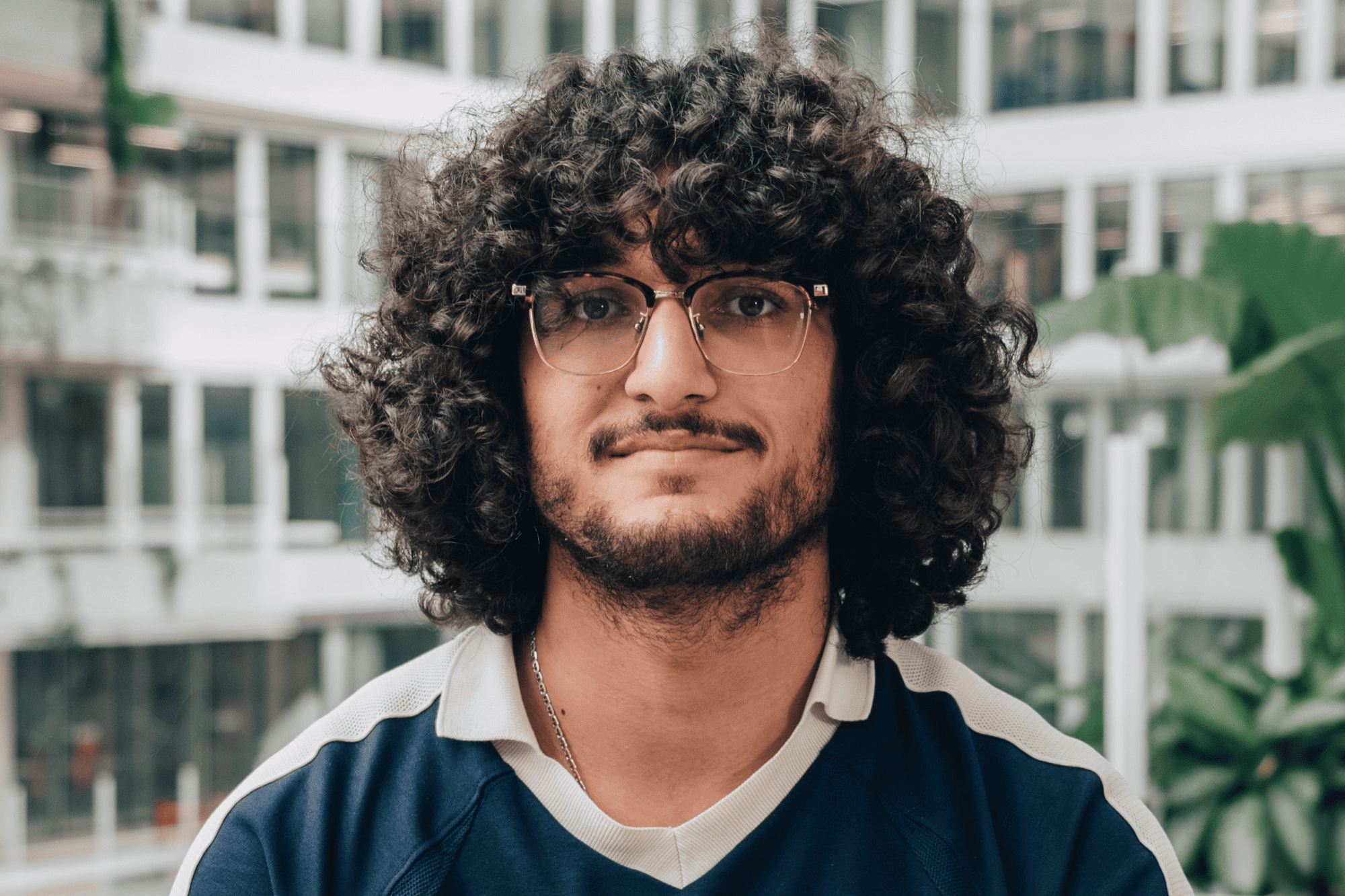 A portrait of Mehdi, an intern, smiling in front of a large, bright office atrium with glass balconies and indoor plants.