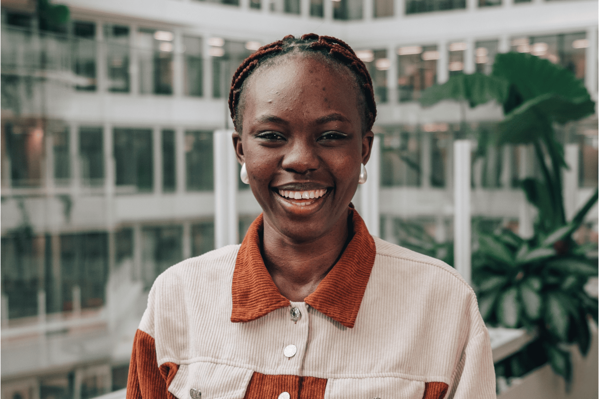 A bright, smiling portrait of Ossene, an intern, wearing a cream and terracotta jacket in a modern glass office atrium.