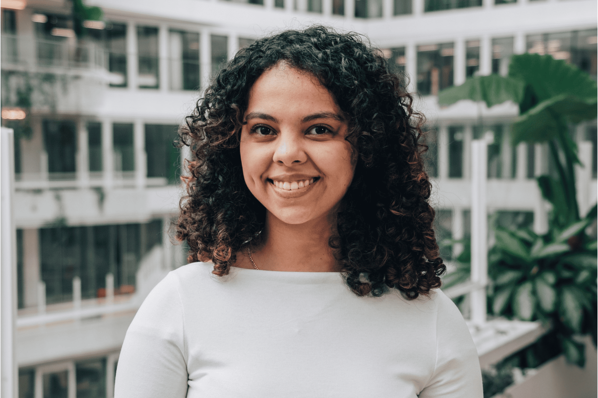 Racha smiling warmly in a white top, standing in a bright, multi-level office environment with greenery.