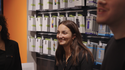 Jenny, a Sales Advisor at an Orange Store, smiling and interacting with colleagues in front of a mobile accessory display.