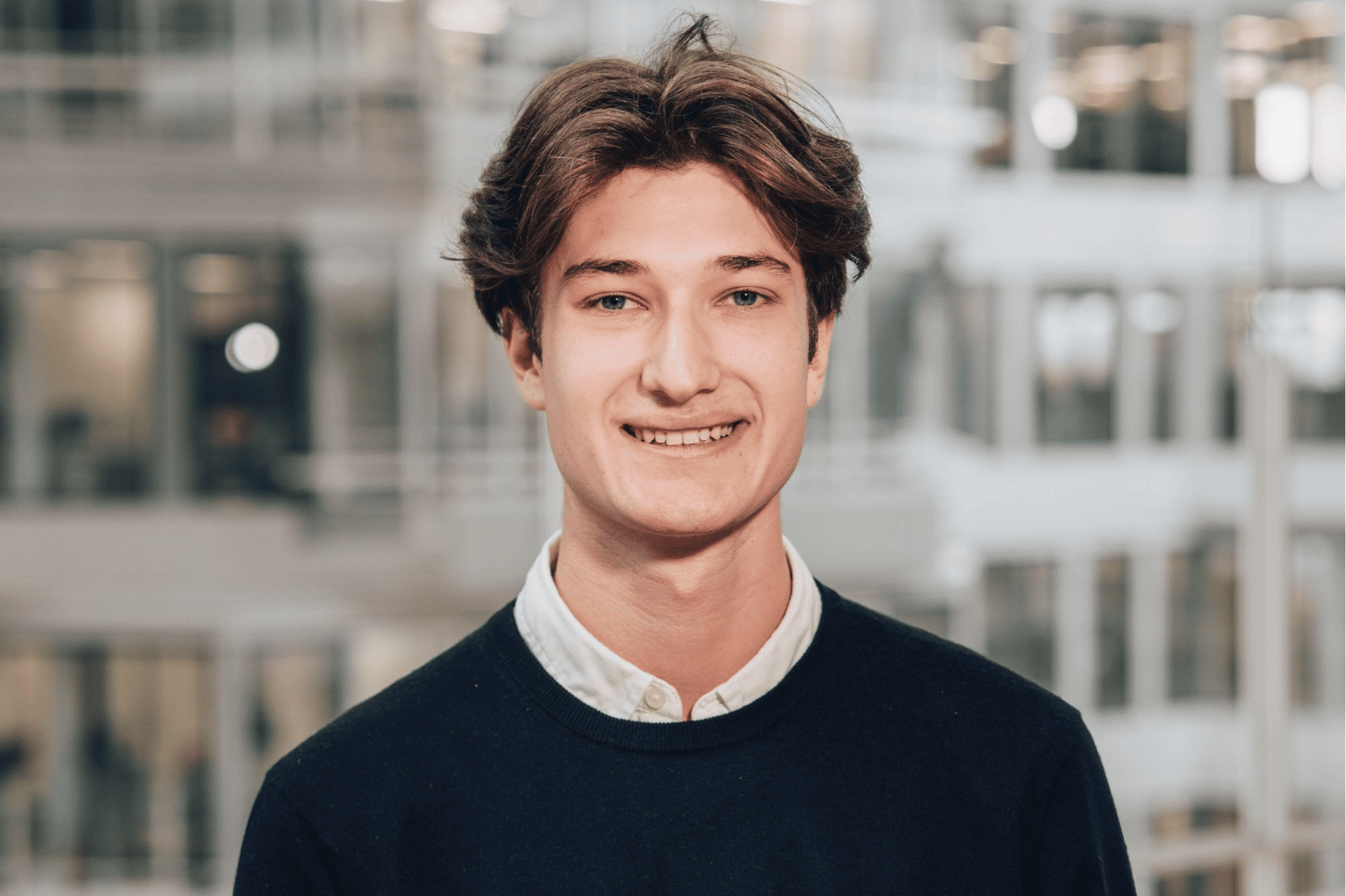 A professional headshot of Clement, a student intern, smiling in a modern office building with a blurred architectural background.