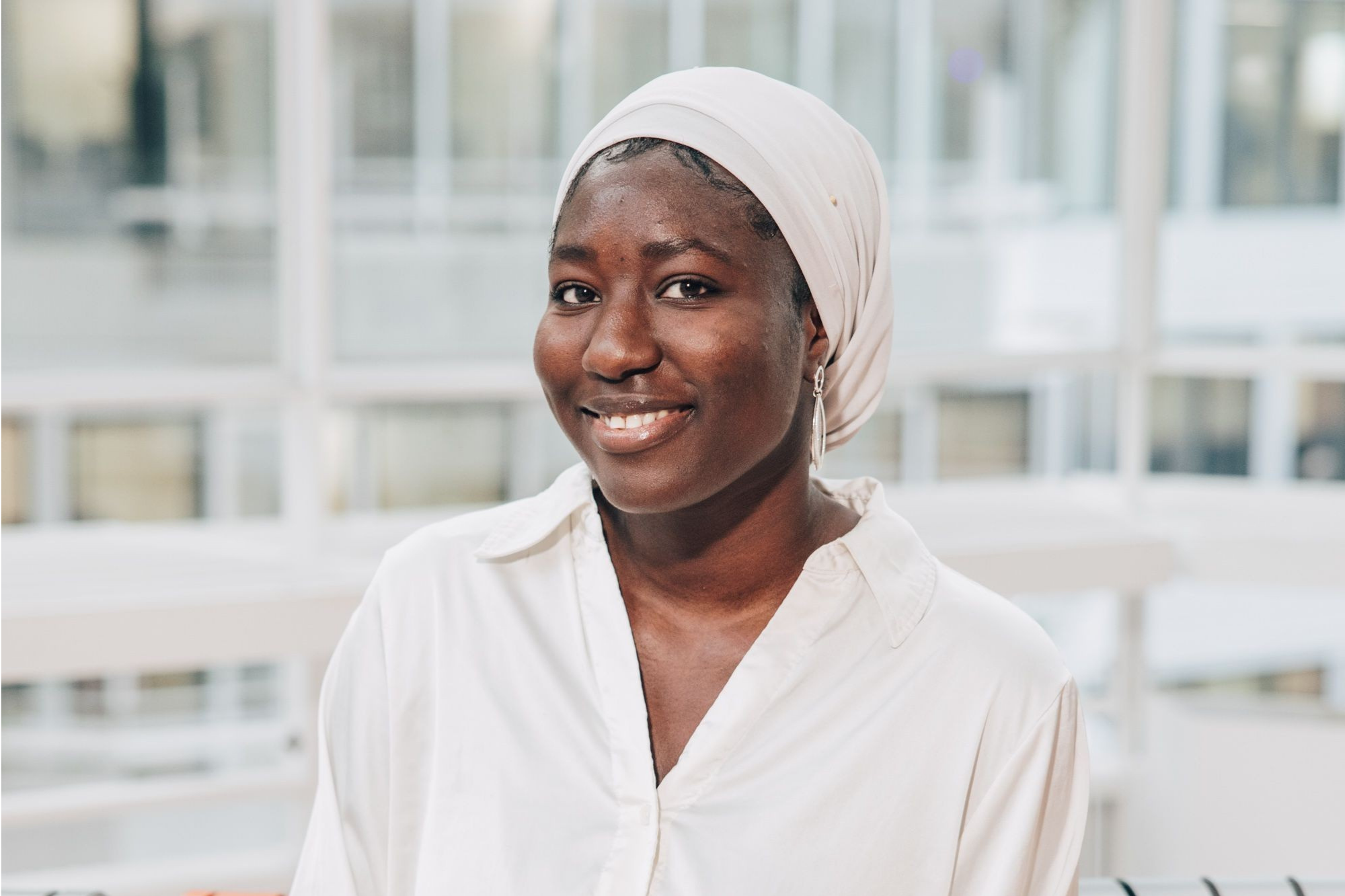 A friendly portrait of Ndeyelyka, an intern, wearing a white headscarf and shirt, smiling in a light-filled professional workspace.