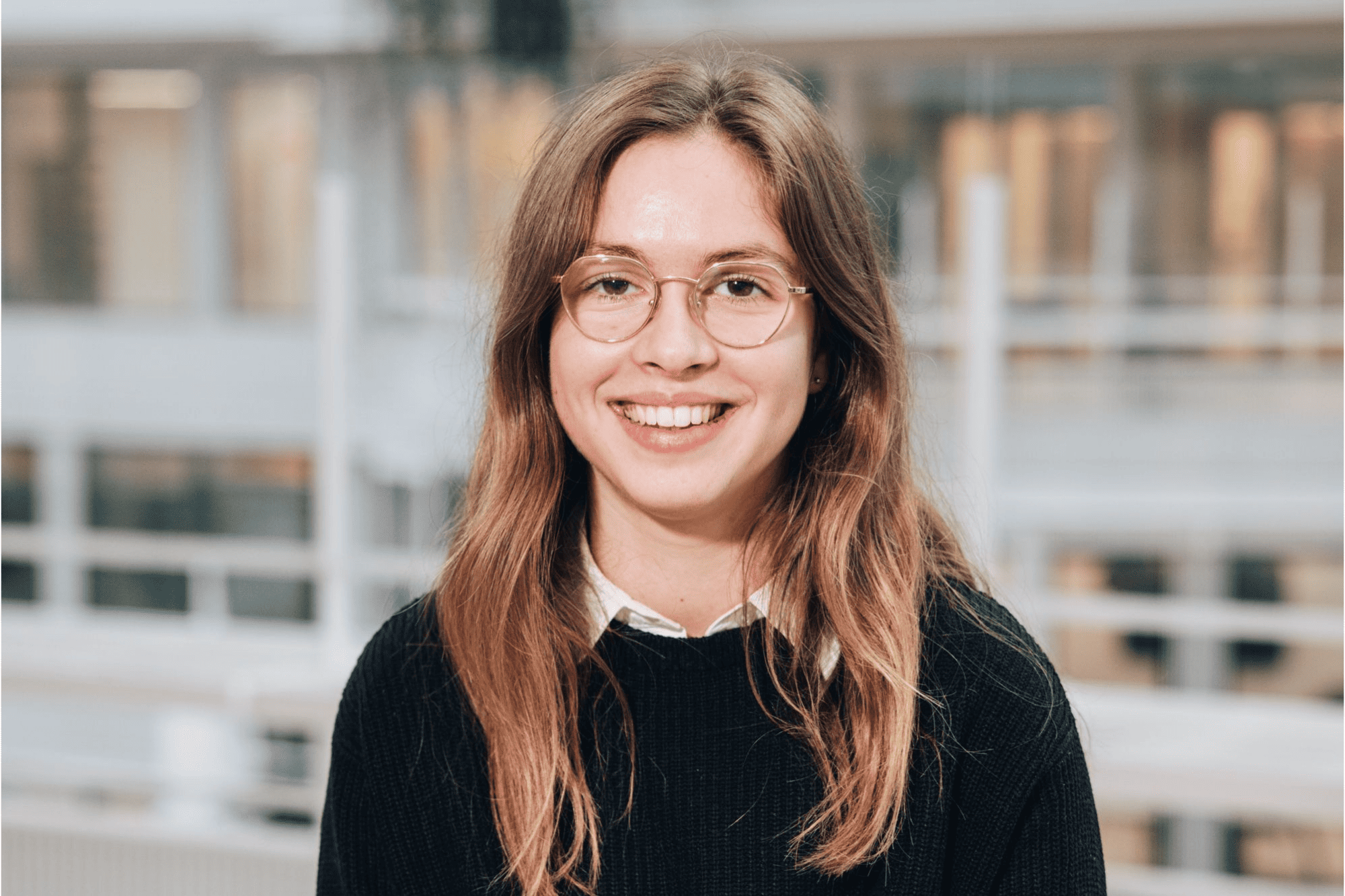 Victoire, a work-study student with glasses and a black sweater, smiling in a modern, open-plan office setting.