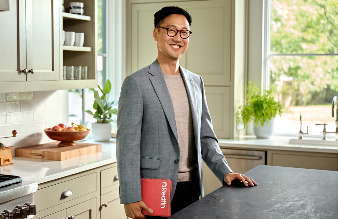 Smiling real estate agent holding a Redfin folder, standing in a bright, modern kitchen with light cabinets and plants.